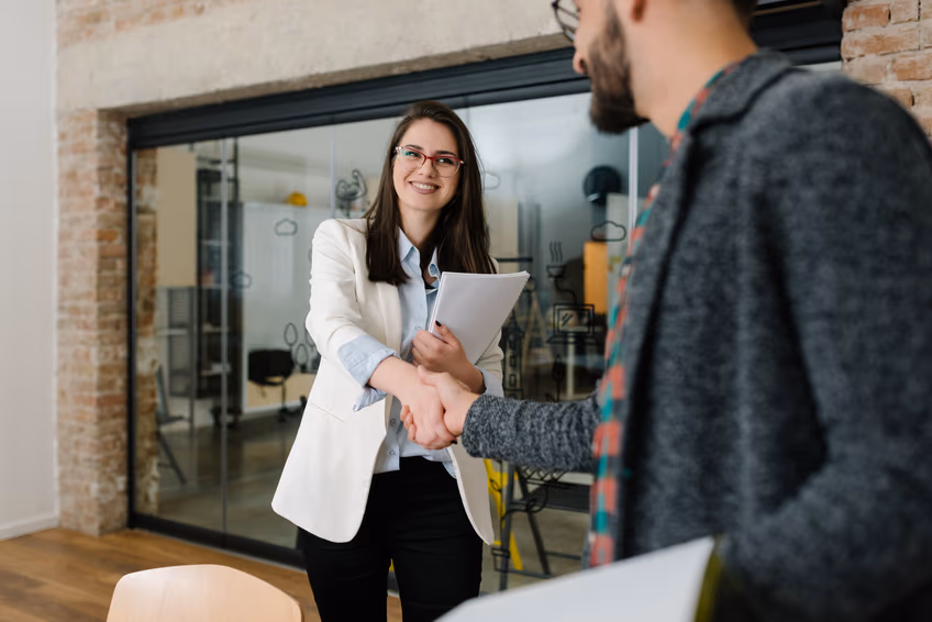 Two people shaking hands at the start of a job interview in an office, one of them holding a folder of documents